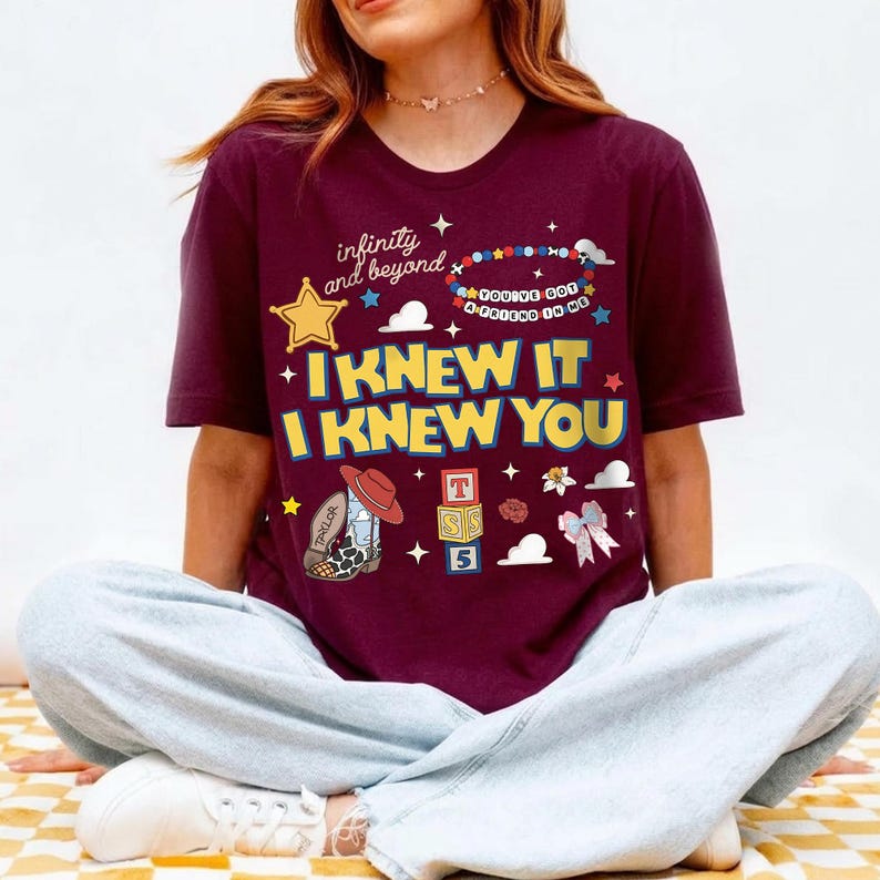 May include: A maroon short-sleeve t-shirt featuring a graphic print with the text "infinity and beyond", "YOU'VE GOT A FRIEND IN ME", "I KNEW IT", and "I KNEW YOU". The design includes a yellow star, a beaded bracelet, a cowboy boot, and blocks.