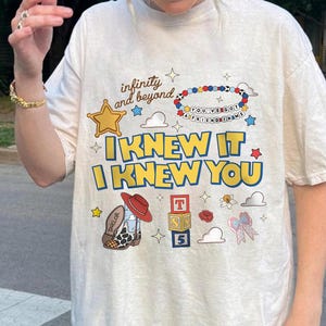 May include: A cream-colored t-shirt featuring a graphic print with the text "infinity and beyond," "I KNEW IT," "I KNEW YOU," and "YOU'VE GOT A FRIEND IN ME." The design includes a beaded bracelet, a cowboy boot, alphabet blocks, and stars.