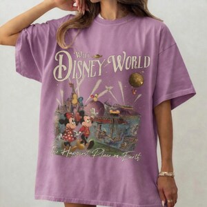 May include: A lilac-colored t-shirt featuring a vintage-style graphic of Walt Disney World. The design includes Mickey and Minnie Mouse, a castle, and the text "The Happiest Place on Earth". The shirt has short sleeves and a relaxed fit.