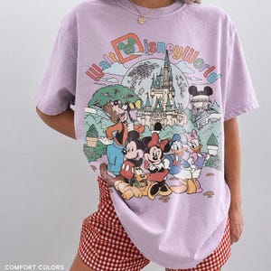 May include: A lilac-colored Walt Disney World graphic t-shirt featuring Mickey Mouse, Minnie Mouse, Donald Duck, Goofy, and Pluto in front of Cinderella's castle. The shirt has the words "Walt Disney World" in a colorful design.