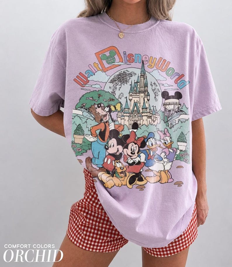 May include: A lilac-colored Walt Disney World graphic t-shirt featuring Mickey Mouse, Minnie Mouse, Donald Duck, Goofy, and Pluto in front of Cinderella's castle. The shirt has the words "Walt Disney World" in a colorful design.