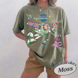 May include: An olive green short-sleeve t-shirt featuring a graphic of Buzz Lightyear from Space Ranger Spin. The design includes the character flying with wings extended, a laser gun, and small green aliens. The text reads Buzz Lightyear's Space Ranger Spin.