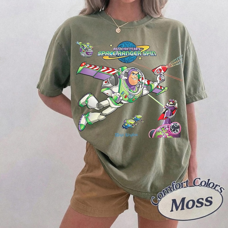 May include: An olive green short-sleeve t-shirt featuring a graphic of Buzz Lightyear from Space Ranger Spin. The design includes the character flying with wings extended, a laser gun, and small green aliens. The text reads Buzz Lightyear's Space Ranger Spin.