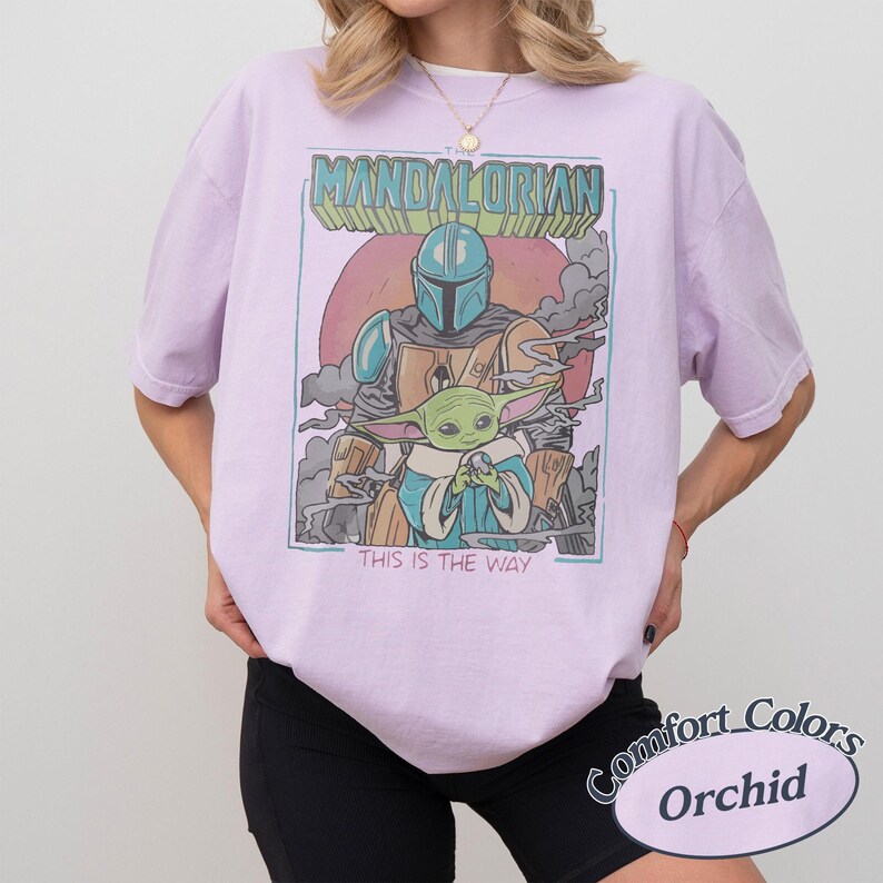 May include: A light purple t-shirt featuring a graphic print of The Mandalorian and Grogu. The design includes the text THE MANDALORIAN at the top and THIS IS THE WAY at the bottom, set against a circular background with smoke elements in muted tones.