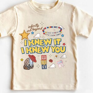 May include: A cream-colored short-sleeve t-shirt featuring a graphic print with the text "infinity and beyond," "YOU'VE GOT A FRIEND IN ME," and "I KNEW IT I KNEW YOU." The design includes a sheriff star, a beaded bracelet, a cowboy boot, blocks, and a bow.
