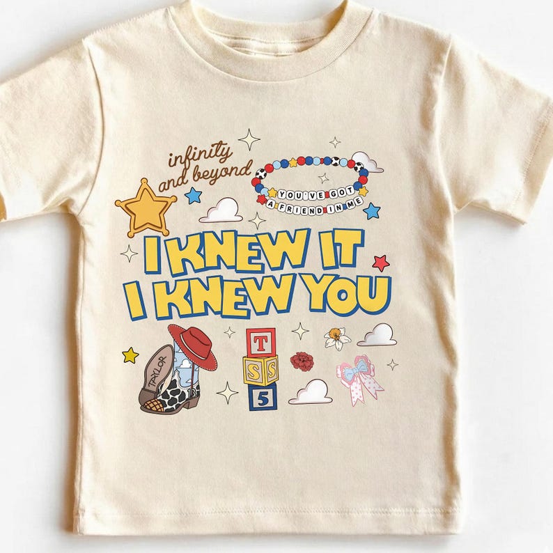 May include: A cream-colored short-sleeve t-shirt featuring a graphic print with the text "infinity and beyond," "YOU'VE GOT A FRIEND IN ME," and "I KNEW IT I KNEW YOU." The design includes a sheriff star, a beaded bracelet, a cowboy boot, blocks, and a bow.