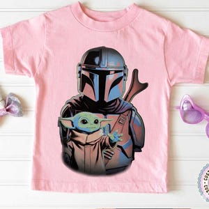 K&ouml;nnte beinhalten: Ein hellrosa T-Shirt mit kurzen &Auml;rmeln und einem Grafikdruck, der eine gepanzerte Figur mit dunklem Helm hinter einem kleinen gr&uuml;nen Wesen mit gro&szlig;en Ohren und Augen zeigt. Die Figur tr&auml;gt einen dunklen Anzug und das Wesen eine beige Robe.