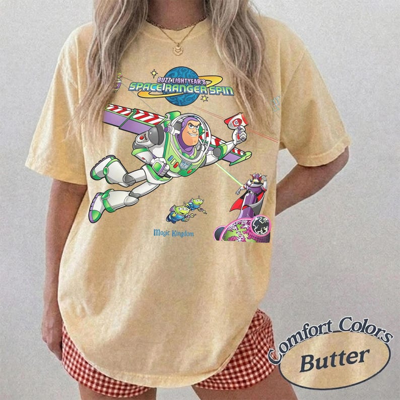 May include: A pale yellow short-sleeve t-shirt featuring a graphic of Buzz Lightyear flying with wings extended. The design includes the text Buzz Lightyear Space Ranger Spin and Magic Kingdom, along with small green alien characters and Zurg.