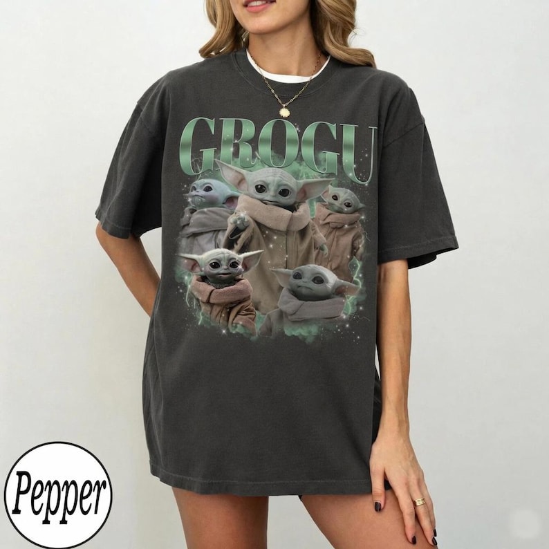 May include: Dark gray t-shirt featuring a graphic of Grogu, also known as Baby Yoda, from The Mandalorian. The design includes the word "GROGU" in green above multiple images of the character. The shirt is a relaxed fit.