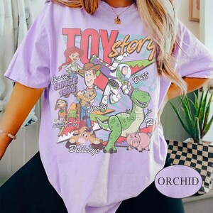 May include: A light purple short-sleeve t-shirt featuring a graphic print of Toy Story characters including Woody, Buzz Lightyear, Jessie, Rex, Slinky Dog, and Bullseye. The text reads Toy Story, Jessie, Buzz, Woody, Slinky Dog, Bullseye, and Since 1995.