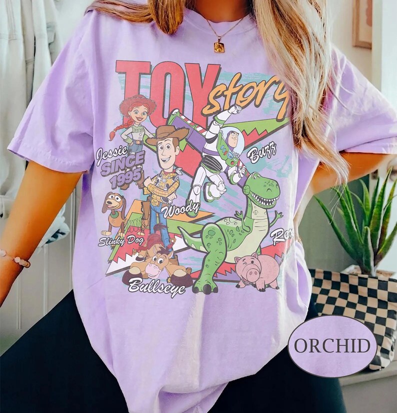 May include: A light purple short-sleeve t-shirt featuring a graphic print of Toy Story characters including Woody, Buzz Lightyear, Jessie, Rex, Slinky Dog, and Bullseye. The text reads Toy Story, Jessie, Buzz, Woody, Slinky Dog, Bullseye, and Since 1995.
