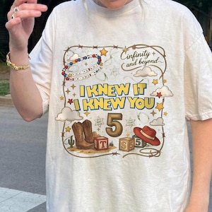 May include: A cream-colored t-shirt featuring a graphic print with the text "I KNEW IT I KNEW YOU", "5", and "Cinfinity and beyond". The design includes cowboy boots, a red hat, alphabet blocks, stars, clouds, and a lasso border.