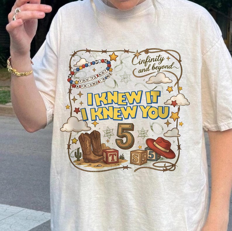May include: A cream-colored t-shirt featuring a graphic print with the text "I KNEW IT I KNEW YOU", "5", and "Cinfinity and beyond". The design includes cowboy boots, a red hat, alphabet blocks, stars, clouds, and a lasso border.