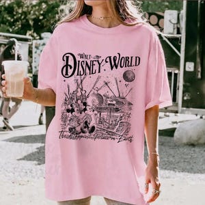 May include: A light pink t-shirt featuring a black graphic of Walt Disney World with Mickey and Minnie Mouse. The text "Walt Disney World" is arched across the top, and "The Happiest Place on Earth" is printed below the graphic.