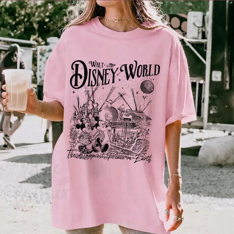 May include: A light pink t-shirt featuring a black graphic of Walt Disney World with Mickey and Minnie Mouse. The text "Walt Disney World" is arched across the top, and "The Happiest Place on Earth" is printed below the graphic.