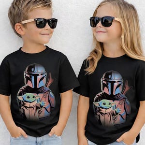 K&ouml;nnte beinhalten: Ein schwarzes Kurzarm-T-Shirt mit einem Grafikdruck eines behelmten Charakters in silberner und blauer R&uuml;stung, der hinter einer kleinen gr&uuml;nen Kreatur mit gro&szlig;en Ohren steht. Das Motiv ist mittig auf der Brust des Kleidungsst&uuml;cks platziert.