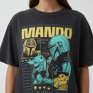 以下が含まれることがあります： マンダロリアンのマンダとグローグのグラフィックが特徴の濃い灰色のTシャツ。デザインには、黄色の「MANDO」とティールの「GROGU」の文字、キャラクターの画像、そして「Wherever I go, he goes」というフレーズが含まれています。