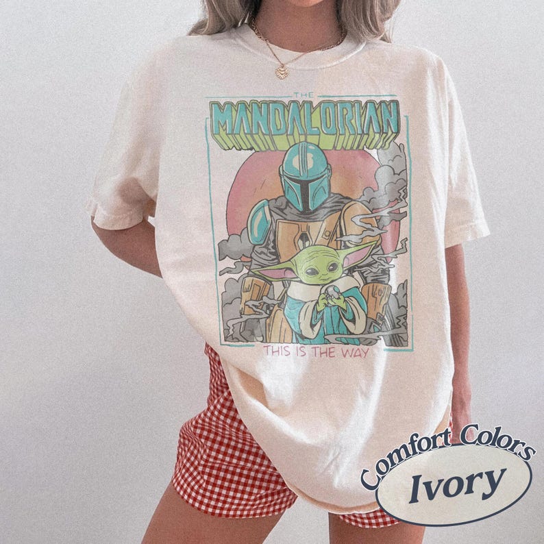 May include: An ivory short-sleeve t-shirt featuring a graphic print of The Mandalorian and Grogu. The design includes the text THE MANDALORIAN and THIS IS THE WAY in a retro style with a circular background, smoke elements, and a teal border.