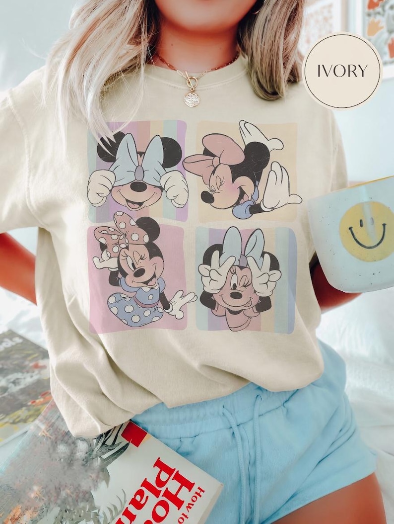 May include: Cream-coloured t-shirt with a four-panel Minnie Mouse graphic. Each panel displays a different pose of Minnie Mouse against a pastel striped backdrop. The shirt is worn with light blue shorts.