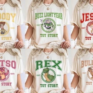 Pu&ograve; includere: T-shirt color crema con personaggi del film "Toy Story". Ogni maglietta mostra il nome di un personaggio in un font universitario, insieme a un'immagine del personaggio e al testo "Toy Story" sottostante. I personaggi includono Woody, Buzz Lightyear, Jessie, Lotso, Rex e Bullseye.