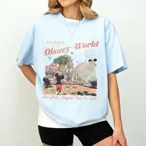 May include: Light blue t-shirt featuring a colorful graphic of Disney World in Florida. The design includes Mickey Mouse, a castle, and the text "Florida Disney World Est. 1971 The Most Magical Place On Earth."