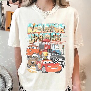 May include: Cream-colored t-shirt featuring a colorful graphic of the town of Radiator Springs with various cartoon cars. The design includes the text "Radiator Springs" and "Lightning McQueen".