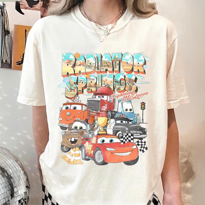 May include: Cream-colored t-shirt featuring a colorful graphic of the town of Radiator Springs with various cartoon cars. The design includes the text "Radiator Springs" and "Lightning McQueen".
