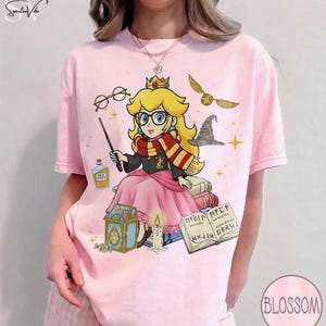 May include: Pink t-shirt featuring a cartoon illustration of Princess Peach as a wizard, wearing glasses and a scarf. The design includes a wand, books, a golden snitch, and a sorting hat. The shirt has the word "Blossom" printed on it.