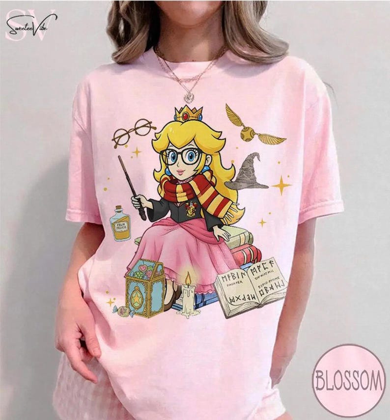 May include: Pink t-shirt featuring a cartoon illustration of Princess Peach as a wizard, wearing glasses and a scarf. The design includes a wand, books, a golden snitch, and a sorting hat. The shirt has the word "Blossom" printed on it.
