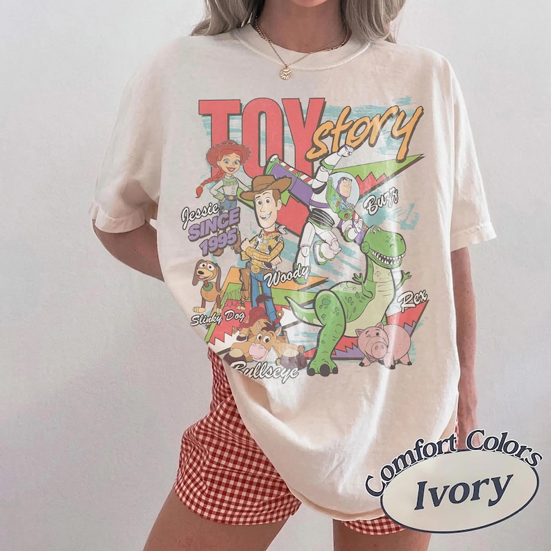 May include: An ivory short-sleeve t-shirt featuring a colorful graphic print of Toy Story characters including Woody, Buzz Lightyear, Jessie, Rex, Slinky Dog, and Bullseye. The text reads Toy Story, Jessie Since 1995, Woody, Buzz, Slinky Dog, and Bullseye.
