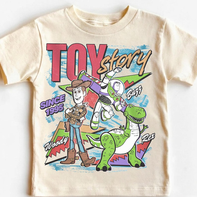 May include: Cream-colored t-shirt featuring the words "TOY STORY" in red and the phrase "SINCE 1995." The shirt has a graphic of Woody, Buzz Lightyear, and Rex the dinosaur. The shirt is a children's apparel item.