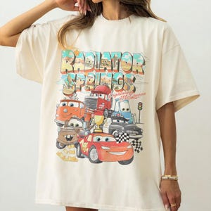 May include: Cream-colored t-shirt featuring a colorful graphic of characters from the movie Cars, including Lightning McQueen. The text "Radiator Springs" is displayed above the cars. The shirt has short sleeves and a relaxed fit.
