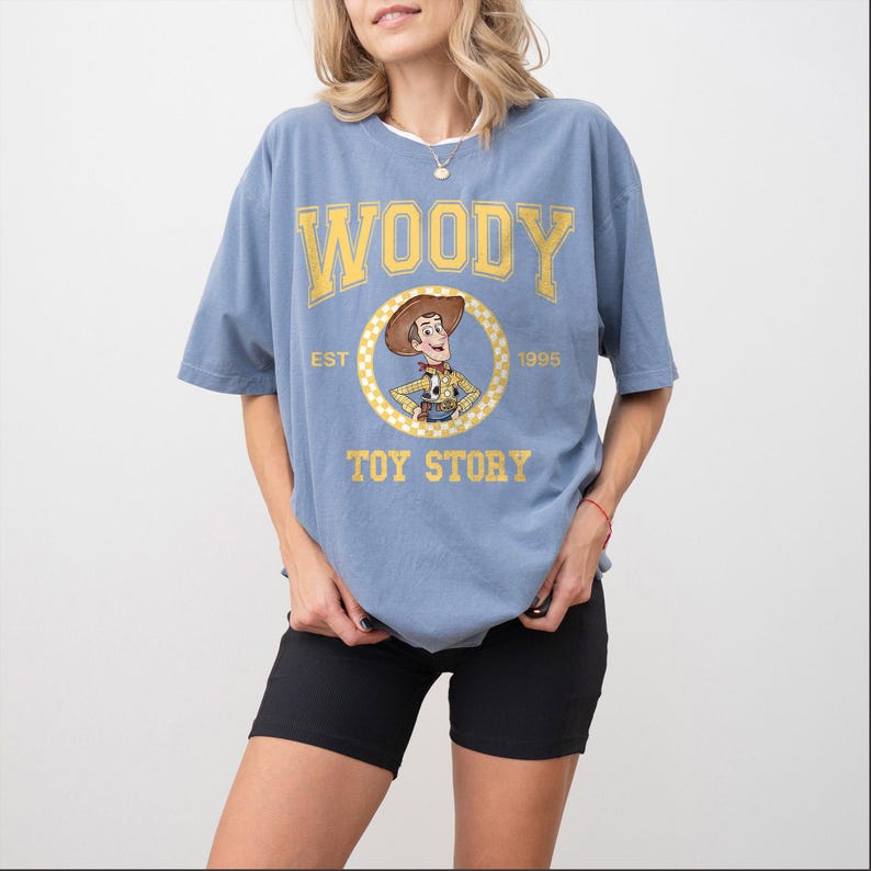 Pu&ograve; includere: T-shirt azzurra con la scritta "WOODY" in lettere gialle. Sotto, una grafica circolare del personaggio Woody di Toy Story. Le parole "EST 1995" e "TOY STORY" sono stampate anche sulla maglietta.