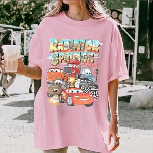 May include: A light pink t-shirt featuring the text "Radiator Springs" in a colorful, retro font. Below the text are cartoon images of cars from the movie Cars. The shirt has short sleeves and a relaxed fit.