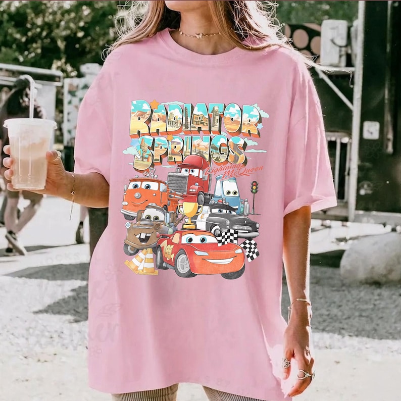 May include: A light pink t-shirt featuring the text "Radiator Springs" in a colorful, retro font. Below the text are cartoon images of cars from the movie Cars. The shirt has short sleeves and a relaxed fit.