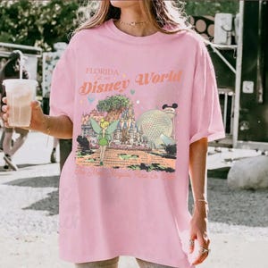 May include: A light pink t-shirt featuring a colorful graphic of Disney World in Florida. The graphic includes Tinkerbell, Cinderella's castle, and Spaceship Earth. Text on the shirt reads "Florida Disney World" and "The Most Magical Place On Earth."