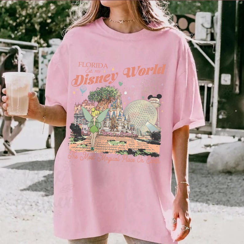 May include: A light pink t-shirt featuring a colorful graphic of Disney World in Florida. The graphic includes Tinkerbell, Cinderella's castle, and Spaceship Earth. Text on the shirt reads "Florida Disney World" and "The Most Magical Place On Earth."