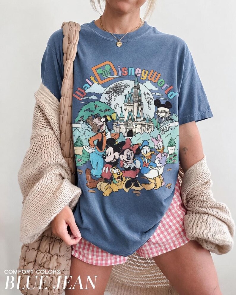May include: Blue jean colored t-shirt featuring a colorful graphic of Disney World characters and the park's logo. The shirt has a vintage look and feel. The words "COMFORT COLORS" and "BLUE JEAN" are printed at the bottom of the image.