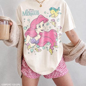 May include: An ivory short-sleeve t-shirt featuring a graphic print of Ariel from The Little Mermaid with Flounder and seahorses, surrounded by light blue bubbles. The text Disney The Little Mermaid is printed in blue above the character.