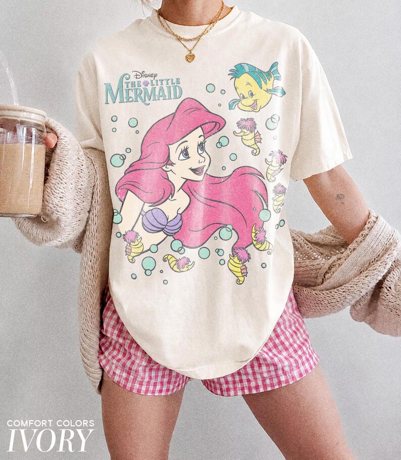 May include: An ivory short-sleeve t-shirt featuring a graphic print of Ariel from The Little Mermaid with Flounder and seahorses, surrounded by light blue bubbles. The text Disney The Little Mermaid is printed in blue above the character.