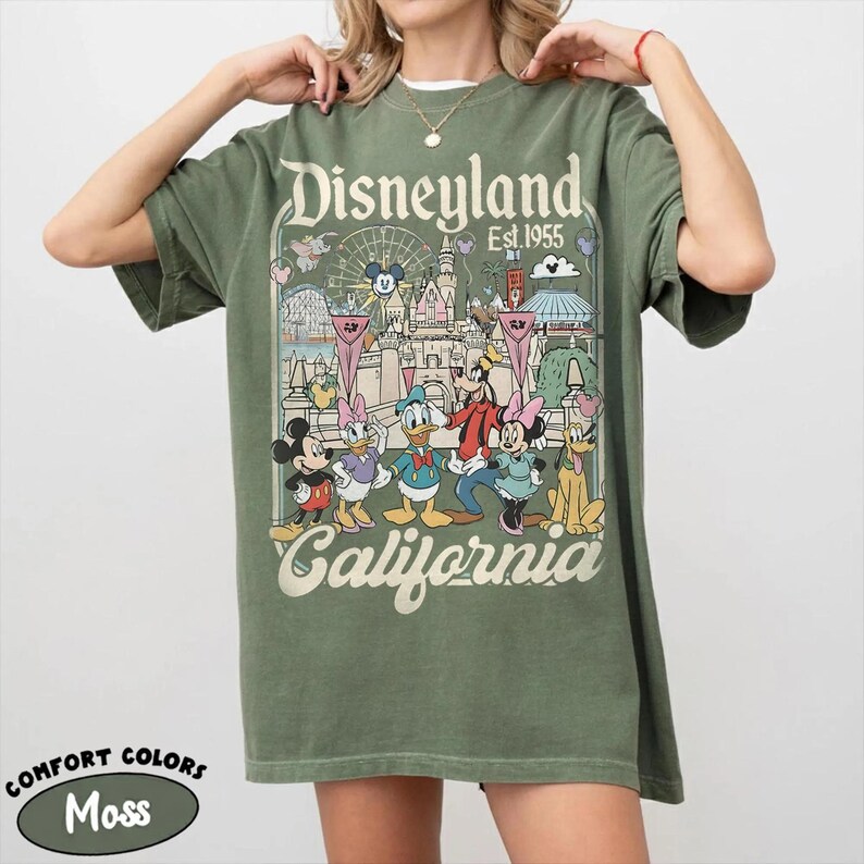 May include: Moss green t-shirt featuring a Disneyland graphic with Mickey Mouse, Donald Duck, and other Disney characters. The shirt has the words "Disneyland California Est. 1955" printed on it. The shirt is a Comfort Colors brand.