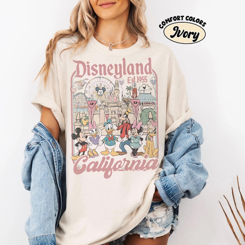 May include: Ivory-colored t-shirt featuring a Disneyland graphic. The design includes the Disneyland logo, the phrase "Est 1955," and various Disney characters. The shirt is part of the Comfort Colors collection.