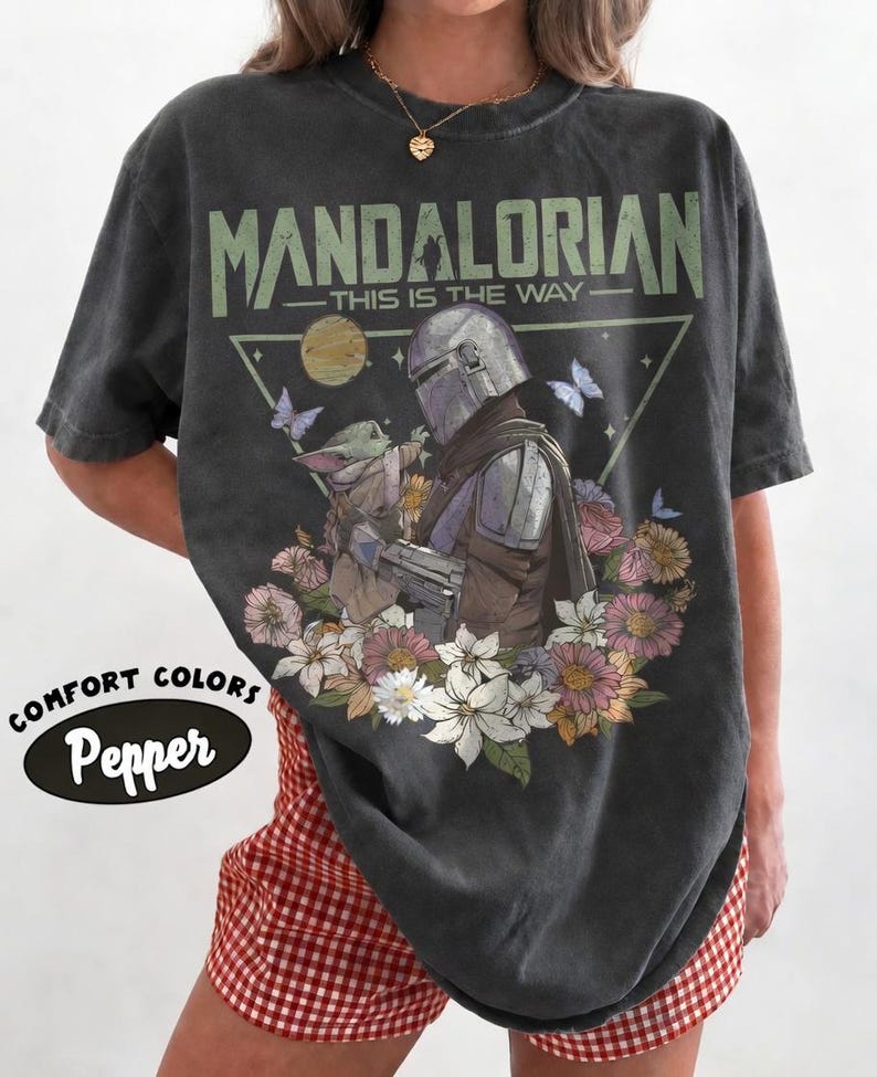 May include: Dark gray t-shirt featuring a graphic of The Mandalorian and Grogu surrounded by flowers and butterflies. The text "MANDALORIAN - THIS IS THE WAY" is printed above the image. The shirt has a Comfort Colors Pepper label.
