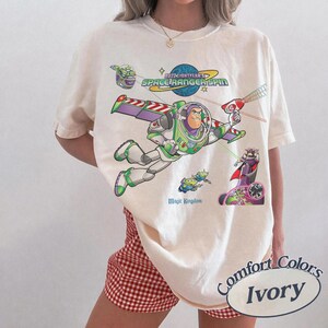 May include: An ivory t-shirt featuring a graphic print of Buzz Lightyear from Space Ranger Spin. The design includes Buzz flying with a laser, Zurg, and small green aliens. The text reads Buzz Lightyear's Space Ranger Spin and Magic Kingdom.