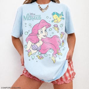May include: A light blue t-shirt featuring a graphic print of Ariel from Disney's The Little Mermaid, along with Flounder and several seahorses surrounded by bubbles. The text Disney The Little Mermaid is printed in the upper left corner.