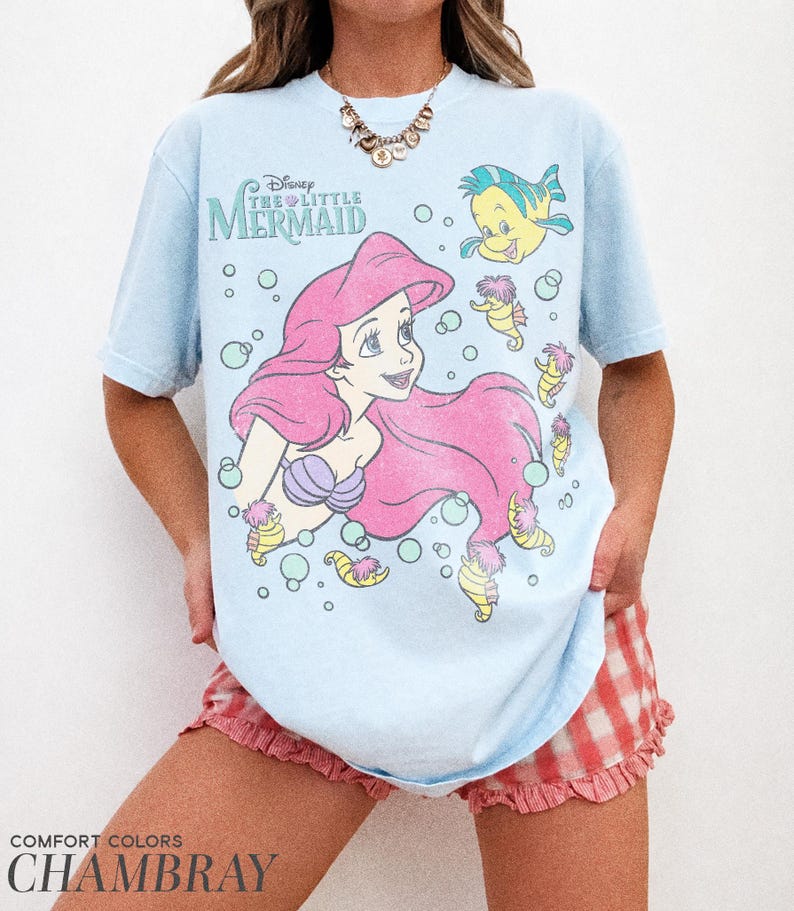 May include: A light blue t-shirt featuring a graphic print of Ariel from Disney's The Little Mermaid, along with Flounder and several seahorses surrounded by bubbles. The text Disney The Little Mermaid is printed in the upper left corner.