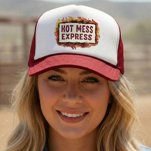 May include: A trucker hat featuring a white front panel with a graphic of a rectangular sign surrounded by flames. The text HOT MESS EXPRESS is printed in dark red letters inside the sign. The brim and mesh back panels are a deep maroon color.