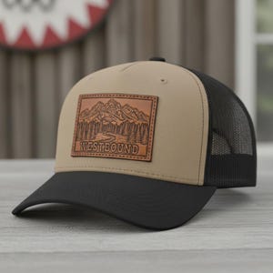 Könnte beinhalten: Beige-schwarze Baseballkappe mit Mesh-Rückseite. Auf der Vorderseite befindet sich ein brauner Lederaufnäher mit einer Berg- und Waldszene und dem Wort "WESTBOUND".
