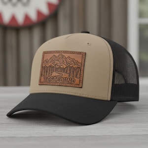 Op de afbeelding: Een beige en zwarte baseballcap met een mesh achterkant. De voorkant heeft een bruin leren patch met een berg- en boslandschap en het woord "WESTBOUND".