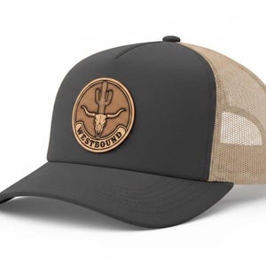 May include: A black trucker hat with a tan mesh back and a curved brim. The hat features a circular leather patch with a cactus and bull skull design, and the word "WESTBOUND" is printed on the patch.